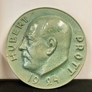Das Bild zeigt ein grün glasiertes Kopfrelief mit der Aufschrift "Hubert Prott 1924"