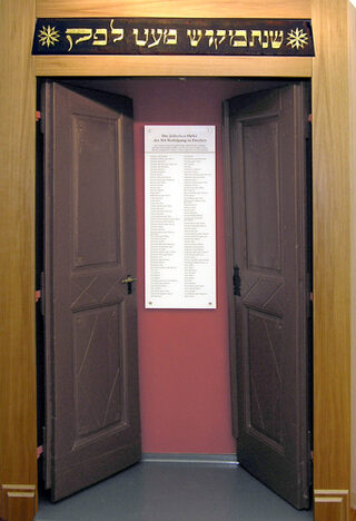 Das Bild zeigt die restaurierte Türe der ehemaligen Frechener Synagoge. Diese wurde an der Wand befestigt, dahinter befindet sich eine weiße Tafel auf roter Wand mit den Namen der jüdischen Opfer der NS-Verfolgung.